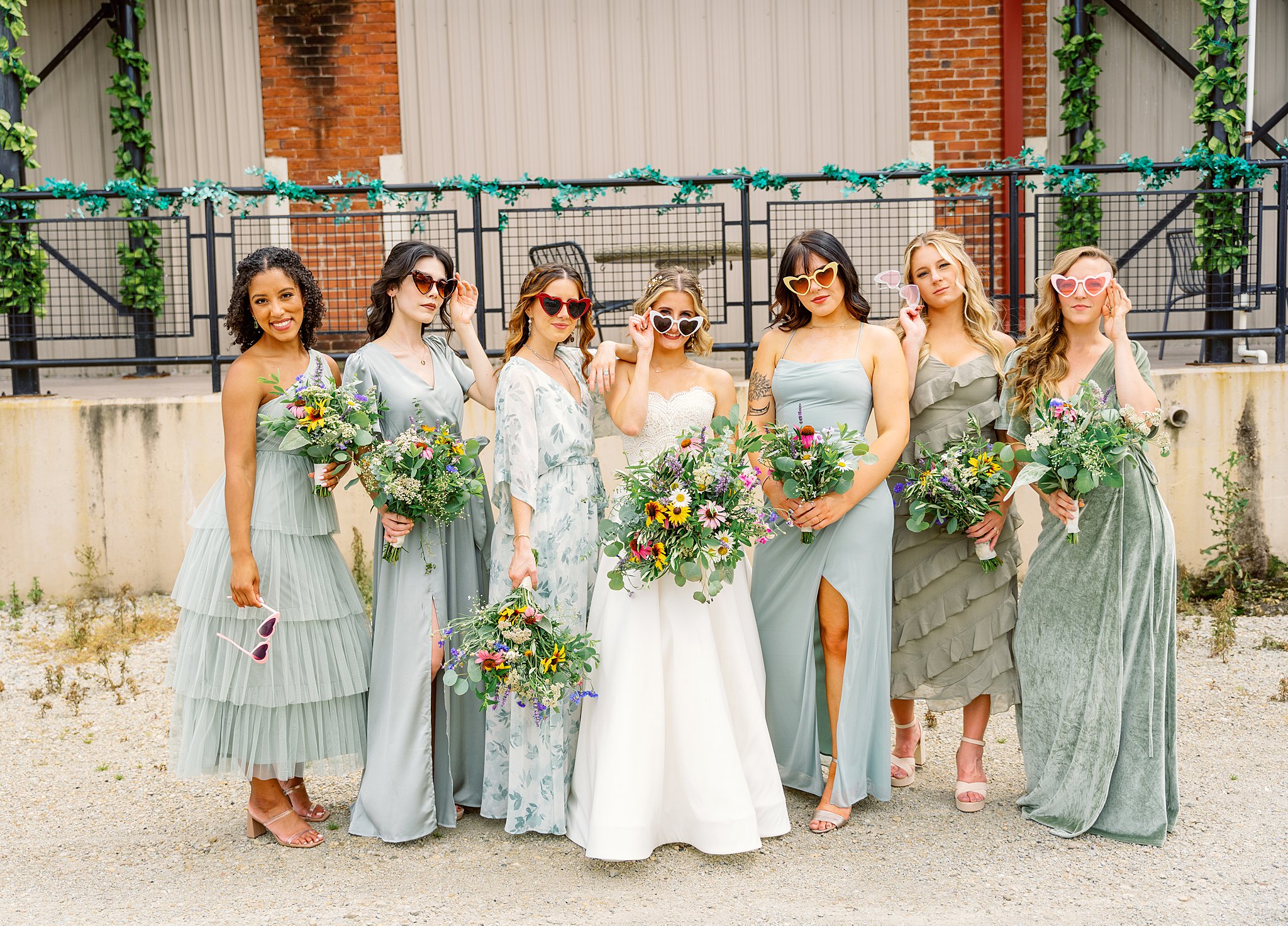 Bridal party in green dresses and heart shaped sunglasses stand outside a modern industrial building unique wedding venues dayton ohio