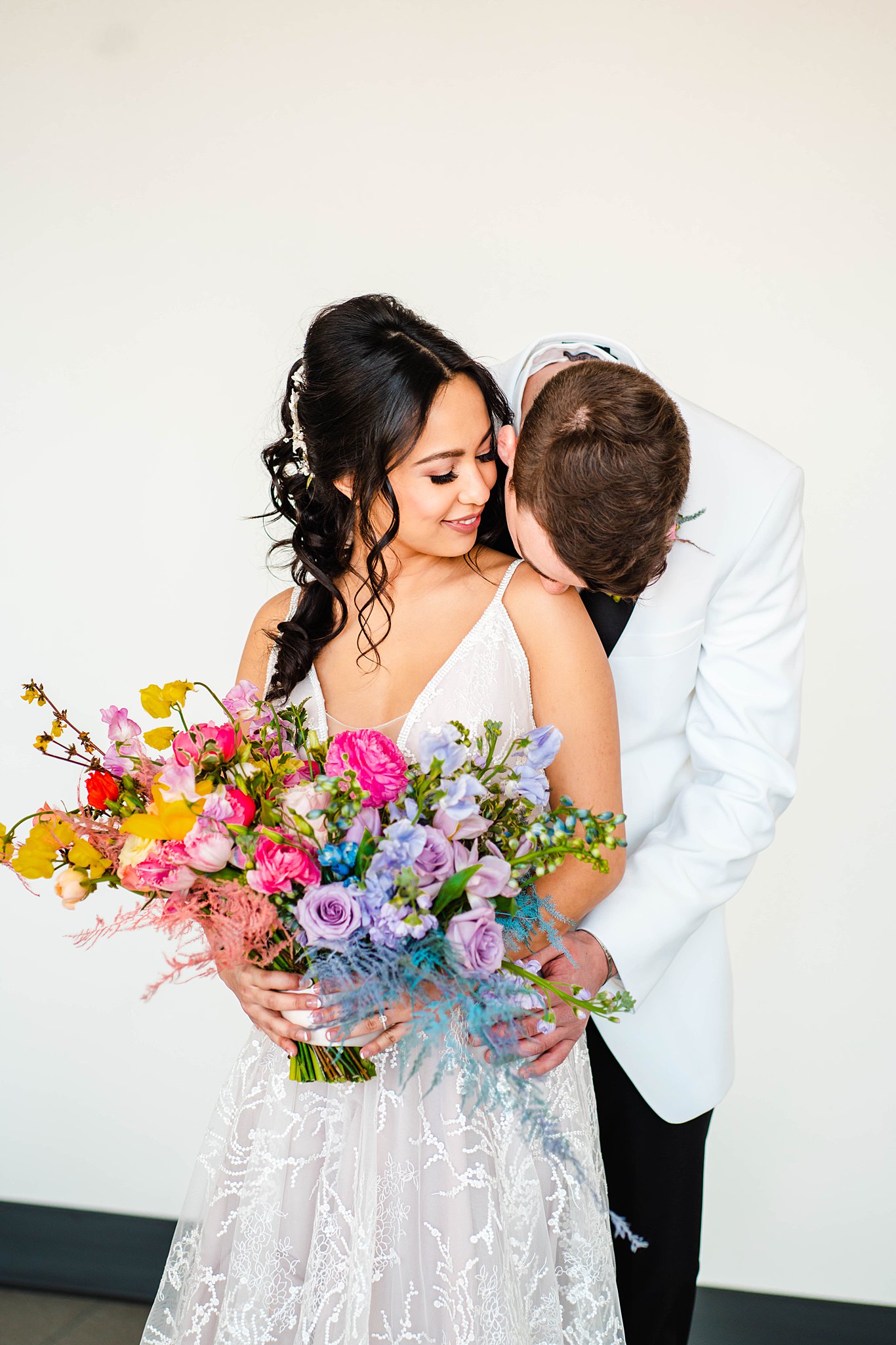 Grroom in a white suit jacket kisses the shoulder of his bride holding a bright multicolor bouquet unique wedding venues dayton ohio